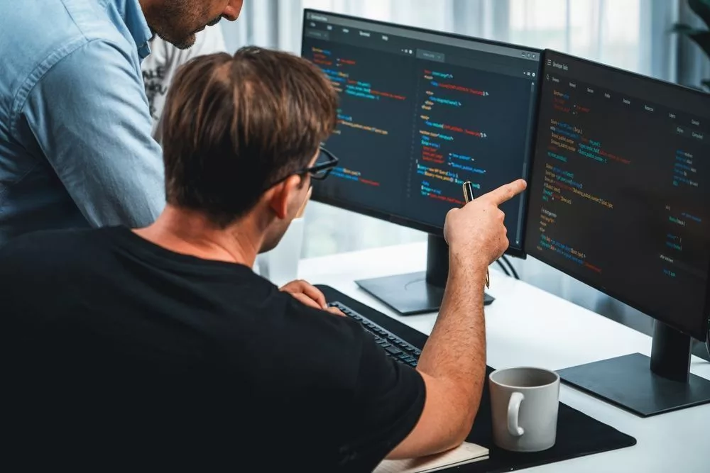 Twee softwareontwikkelaars analyseren code op schermen tijdens overleg